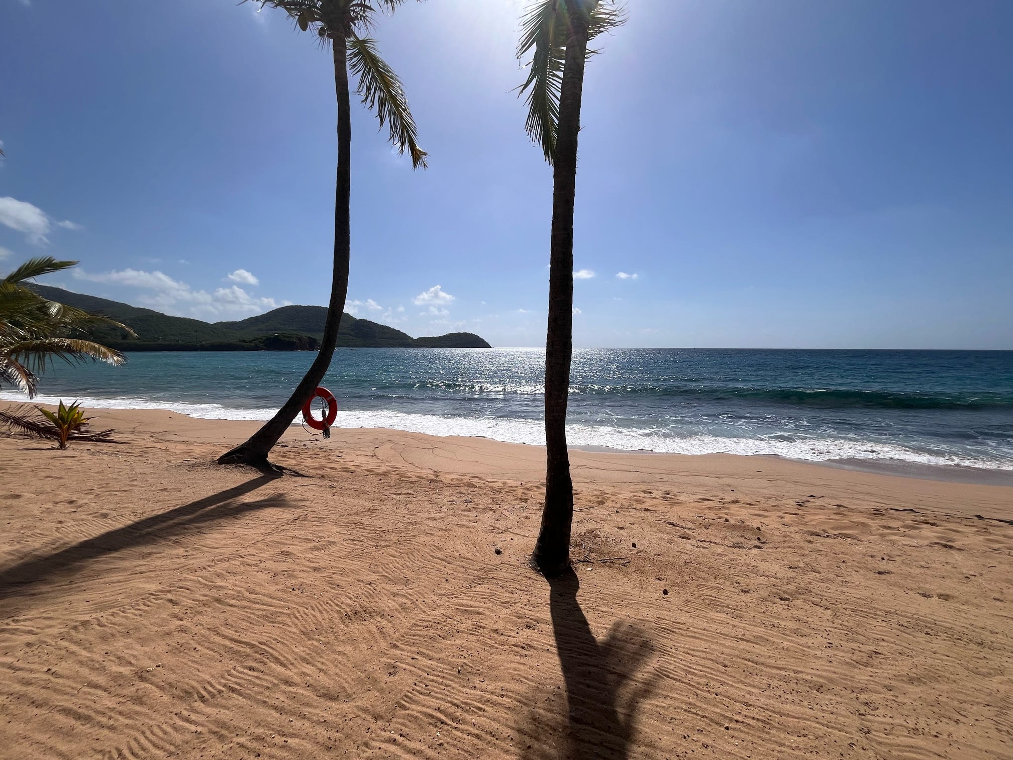 Two palm trees under a full sun on the beach with white surf, a life preserver and the Indian Island in Antigua.